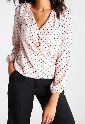 Donna con una camicetta rosa pallido a maniche lunghe con pois neri e pantaloni neri, in piedi con una mano in tasca.