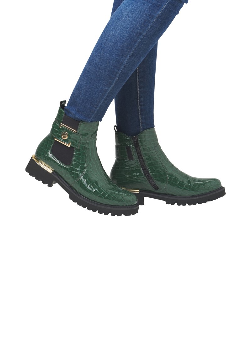 Bottes cheville en cuir vert brillant à texture crocodile avec semelles noires, accents dorés, fermetures éclair latérales et boucle dorée, portées avec un jean bleu.