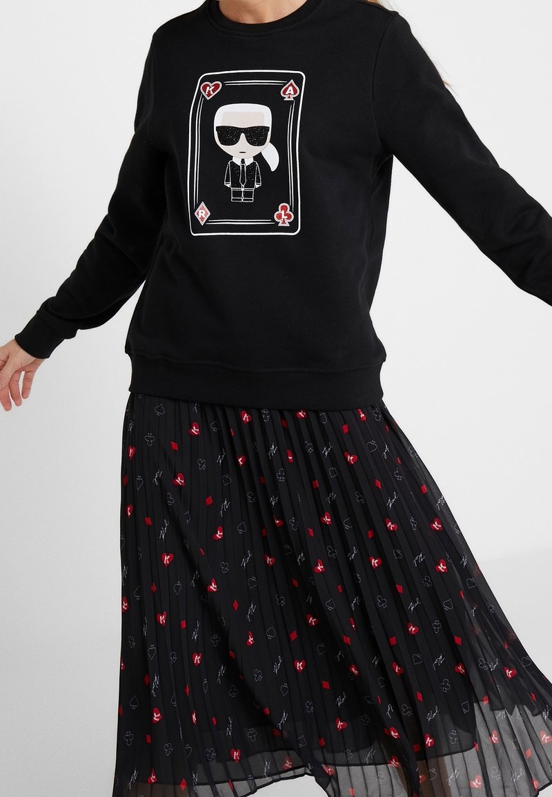 Sweat-shirt noir avec un motif de carte montrant une silhouette stylisée portant des lunettes de soleil, associé à une jupe plissée noire ornée de figures de cartes rouges et blanches.