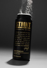 Svart cylindrisk behållare med STMTNT vaxpulver, med guldig text. Locket är öppet och pulver rinner ut, mot en mörk bakgrund.