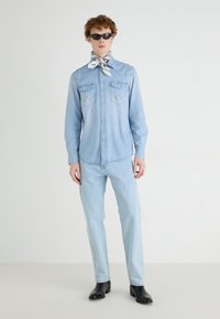 Camisa de denim azul claro con dos bolsillos en el pecho y una yugo puntiagudo, combinada con unos pantalones vaqueros azul claro a juego y botas negras hasta el tobillo, terminada con una bufanda estampada.