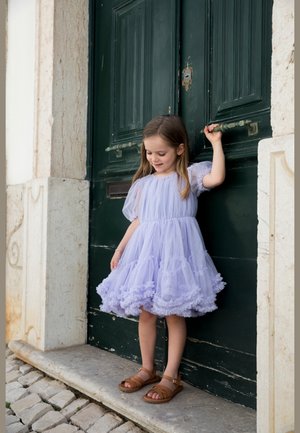 Pige i lyse lilla kjole og brune sandaler, der holder i håndtaget på en mørkegrøn dør, stående på en stentrin ved brosten.