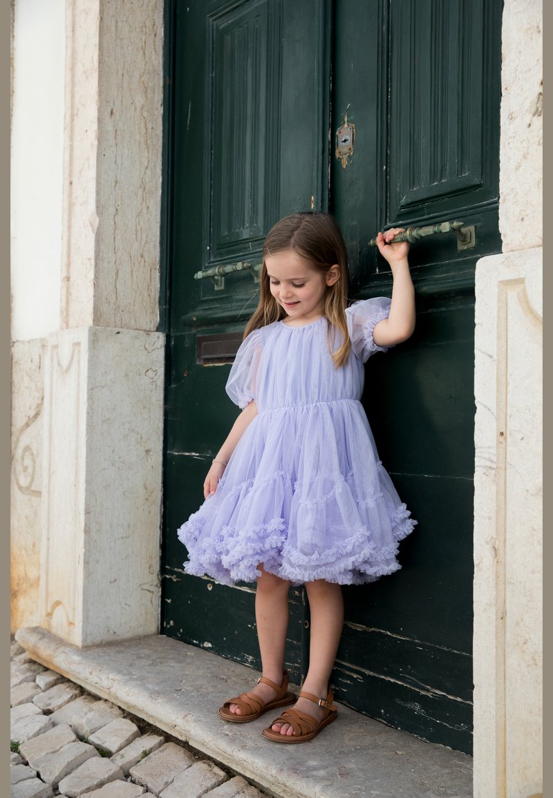 Κορίτσι με ανοιχτό μωβ φόρεμα και καφέ σανδάλια κρατά το χερούλι σκούρας πράσινης πόρτας, στέκεται σε πέτρινο κατώφλι δίπλα σε λιθόστρωτο.