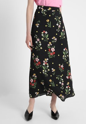 Kvinde iført en sort nederdel til anklerne med flerfarvede blomster- og gule prikmønstre, kombineret med sorte sko og en pink top.