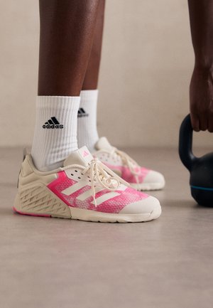 Piernas con calcetines blancos de Adidas y zapatillas rosas de Adidas, de pie sobre un suelo, junto a una mano que sujeta una kettlebell negra.