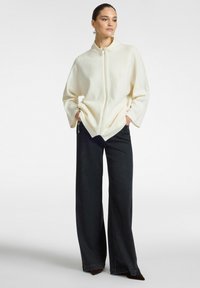 Donna che indossa un ampio maglione bianco con zip e pantaloni neri a gamba larga, in piedi con le mani nelle tasche su uno sfondo neutro.