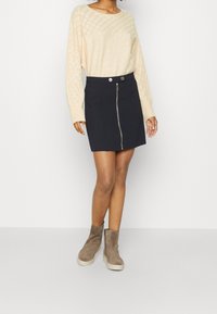 Beige pletený svetr s diamantovým vzorem, doplněný navy mini sukní se zipem vpředu a dvěma kapsami. Hnědé kotníkové boty.