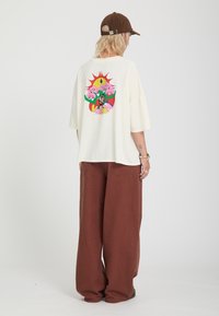 Άτομο που φοράει κρεμ oversized μπλουζάκι με πολύχρωμο γραφικό στην πλάτη, καφέ φαρδιά παντελόνια και καφέ καπέλο μπέιζμπολ, που στέκεται γυρισμένο από την πλάτη.