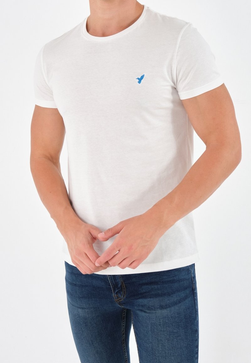 Homme portant un t-shirt blanc uni à manches courtes avec un petit logo d'oiseau bleu sur la poitrine et un jean bleu, debout les mains jointes.