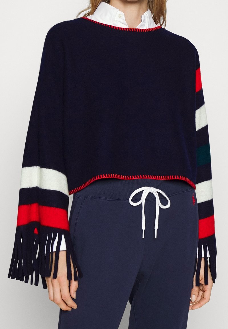 Femme portant un pull court bleu marine avec des rayures rouges, blanches et vertes sur les manches, des détails à franges, associé à un pantalon de survêtement bleu marine et une chemise blanche.