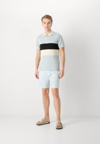 Pólo azul claro com colarinho com fecho, apresentando riscas horizontais em preto, creme e cinza. Combinado com calções risca azul claro e sandálias.