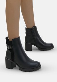 Bottines noires en cuir lisse et en daim, dotées d'un talon bloc, d'une fermeture éclair sur le côté et d'un détail de boucle décoratif. Semelle texturée.