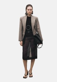 Blazer beige doppiopetto sopra una maglietta nera, abbinato a una gonna midi nera trasparente. Sandali neri e una borsa nera completano il look.