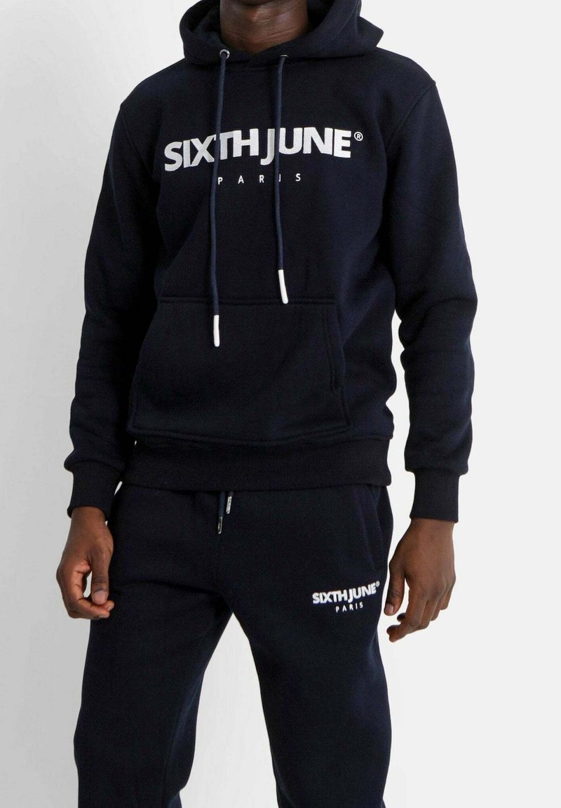 Sweat à capuche bleu marine avec logo blanc "SIXTH JUNE PARIS", capuche à cordon, poche kangourou, pantalon de jogging assorti avec logo sur la jambe. Texture de tissu doux.