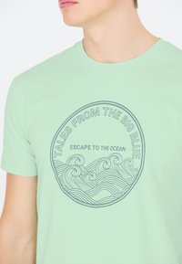 Ljusgrön t-shirt med en cirkulär grafik med vågmotiv och texten "Berättelser från det Stora Blå" och "Flykt till Havet" i grått.