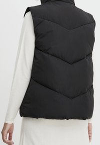 Czarna pikowana kamizelka z wysokim kołnierzem, wzorem przeszyć w kształcie jodełki oraz gładką fakturą. Nosi się ją na jasnym, długim rękawie koszulce.