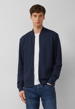 Man met bruin haar die een marineblauw bomberjack draagt over een wit T-shirt en blauwe spijkerbroek, staand tegen een effen grijze achtergrond.