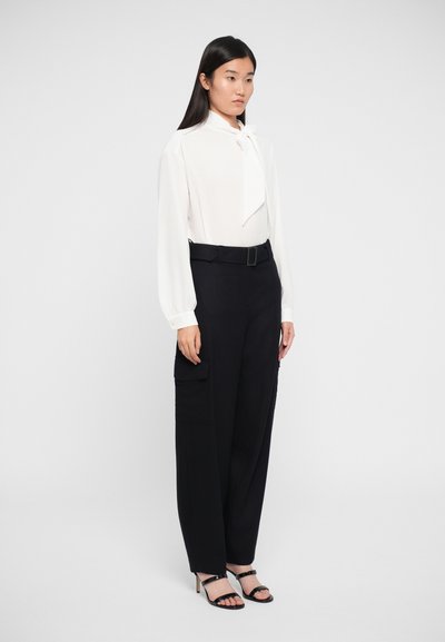 White long-sleeve blouse with bow tie detail, paired with high-waisted black cargo pants and open-toe black heeled sandals. Plain background.