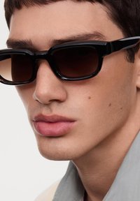 Occhiali da sole neri rettangolari con lenti scure, montatura nera spessa e finitura liscia. Il design presenta angoli angolari.