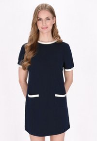 Marineblauwe jurk met korte mouwen, ronde halslijn en crèmekleurige afwerking. Het heeft twee voorzakken met witte accenten. Glad stoftextuur.