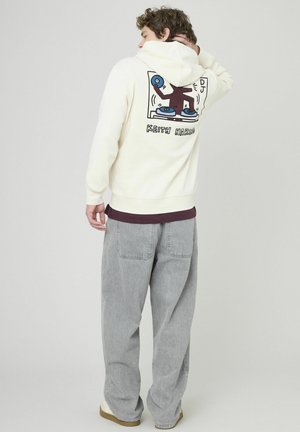 Sweat à capuche crème avec un imprimé graphique au dos, présentant un design de DJ. Assorti à un pantalon baggy gris et des chaussures beiges.