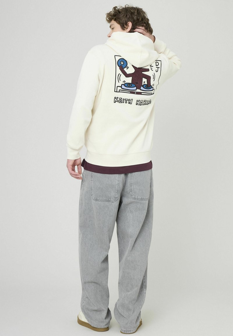 Sweat à capuche crème avec un imprim é graphique au dos, présentant un design de DJ. Assorti à un pantalon baggy gris et des chaussures beiges.