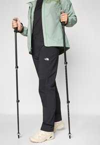 Pantalons de randonnée noirs avec un logo, associés à une veste vert clair, des chaussures beiges et des bâtons de trekking, sur un fond uni.