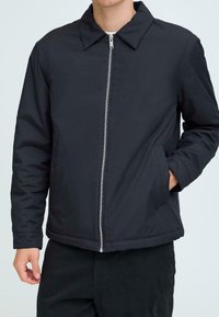 Man wearing a black zip-up jacket with a collar, hand in side pocket, shown from neck to hips against a plain background.