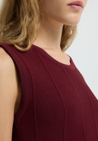 Haut sans manches bordeaux en tissu tricoté texturé. Présente un col rond et des coutures verticales. Coupe structurée, design minimal.