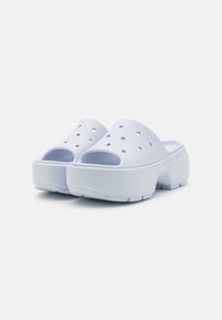Zoccoli bianchi slip-on realizzati in materiale morbido e testurizzato, con fori di ventilazione, una suola piatta spessa e un tacco leggermente sollevato.