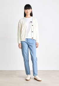 Cardigan crema a treccia con scollo a V e bottoni marroni, abbinato a una maglietta bianca e jeans in denim chiaro a gamba dritta, completato da sneakers crema.