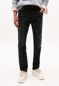 Črne kavbojke iz jeansa z ozkim krojem, ki imajo pet žepov, klasičen gumb za zapiranje in majhno logotipno značko spredaj. Oblečene so z bež supergami.