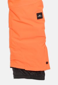 Kirkas oranssit lumihousut mustalla vuorilla, joissa on logopatchi, useita saumoja ja tiukka nilkka turvallisen istuvuuden takaamiseksi.