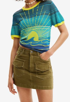 Haut en maille bleu et vert à manches courtes avec un motif jaune de soleil et de vague, porté avec une jupe utilitaire vert olive et un collier de coquillages.