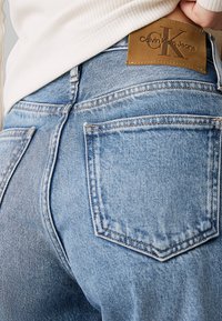 Jeans de mezclilla de un color azul claro que presentan un diseño clásico de cinco bolsillos, con un parche de cuero marrón de Calvin Klein Jeans en la cinturilla.