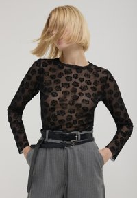 Haut transparent à manches longues noires avec un motif de rose gaufré, col rond, associé à un pantalon gris à rayures tailleur taille haute et une ceinture noire.
