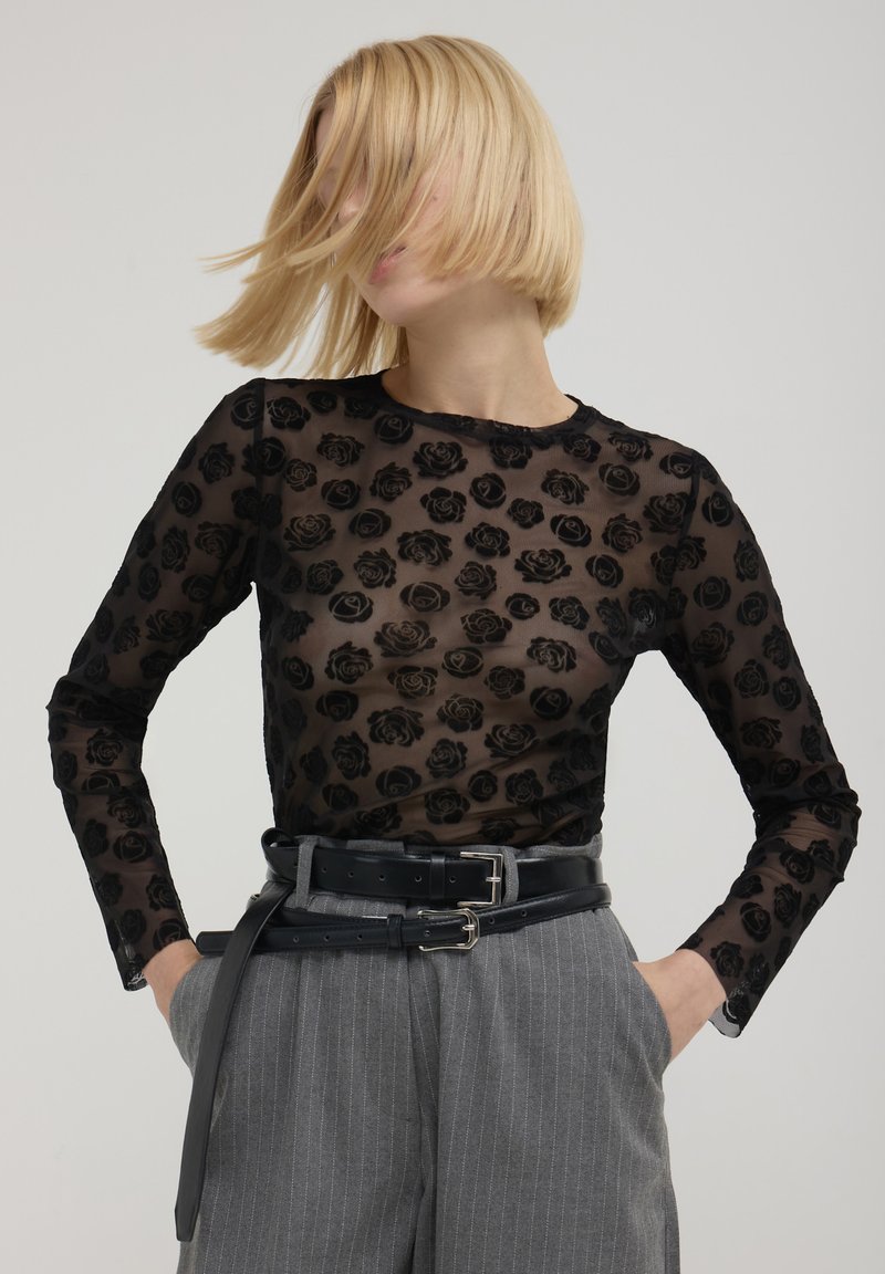 Haut transparent à manches longues noires avec un motif de rose gaufré, col rond, associé à un pantalon gris à rayures tailleur taille haute et une ceinture noire.