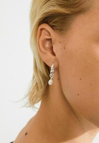 Sølvøreringe med en perledrop, krystalaccentuer og et delikat kædedesign, præsenteret på en modèles øre med kort blond hår.