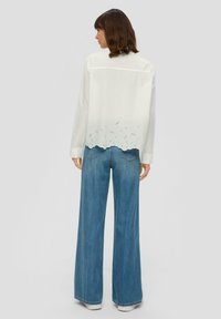 Weiße Bluse mit einer gewellten bestickten Saumkante, kombiniert mit hoch taillierten blauen weitgeschnittenen Jeans. Das Outfit hat lange Ärmel und einen Rückensaum.