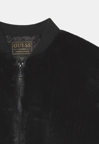 Sort faux fur jakke med ribbet krave og lynlås, med et GUESS-mærke syet indeni nakkeområdet.
