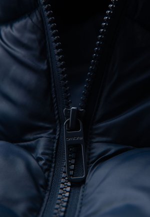 Nahaufnahme einer teilweise zugezippten dunkelblauen gepolsterten Jacke, die den metallischen Reißverschluss mit dem eingravierten Markennamen "ombre" zeigt.