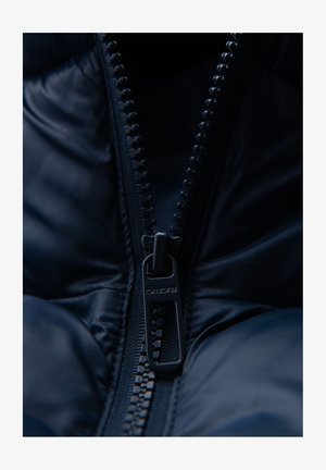 Close-up of a partially zipped dark blue padded jacket showing the metallic zipper pull with the brand name "ombre" engraved.