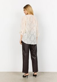 Kvinna vänd bort från kameran iförd en genomskinlig elfenbensfärgad blus av blommig spets med trekvartsärmar, mörka läderbyxor med vida ben och svarta remsandaler med klack.