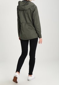 Olivgrön huverjacka med dragsko i midjan, bärs över svarta slitna skinny jeans och vita gympaskor. Bakifrån visas plaggens snitt och passform.