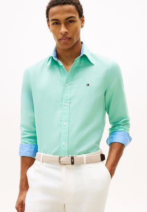Man wearing a mint green button-up shirt with blue rolled sleeves, white pants, and a beige woven belt, standing with one hand in pocket.