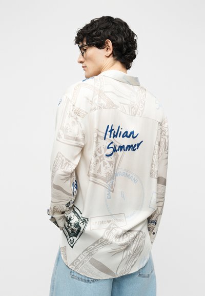 Jeune homme aux cheveux bouclés foncés et lunettes, portant une chemise blanche à motifs avec le texte « Italian Summer », associée à un jean bleu clair, de dos.