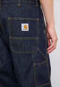 Dark denim overalls featuring yellow stitching, a large back pocket, and a square white label with an orange logo. Close-up of the fabric texture.