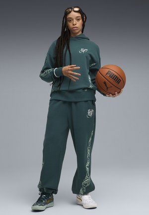 Completo sportivo verde scuro con disegni laterali intricati, maniche lunghe e cappuccio. Tiene un pallone da basket, indossando scarpe colorate con dettagli.