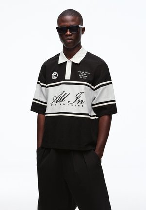 Mann trägt schwarze Sonnenbrille und ein schwarz-weißes Poloshirt mit der Aufschrift "Alles oder Nichts", steht mit den Händen in den Taschen.