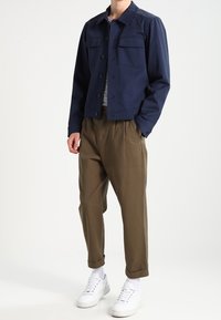 Mörkblå, knäppt jacka med två bröstfickor, bärs över en randig skjorta. Tillsammans med beige, avslappnade byxor och vita sneakers.